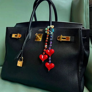 The Big Chakra Energy Bag Charm draped around the handle of a Hermès Birkin bag, with its vibrant chakra gemstones adding a playful, colorful contrast to the luxury leather accessory.