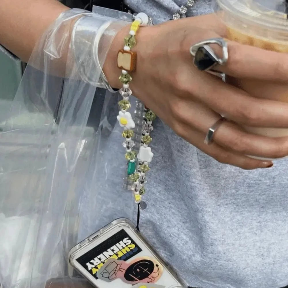 Person holding an iced coffee with a phone hanging from their wrist by the Breakfast Club Wristlet Phone Strap, showcasing colorful breakfast-themed beads in natural light.